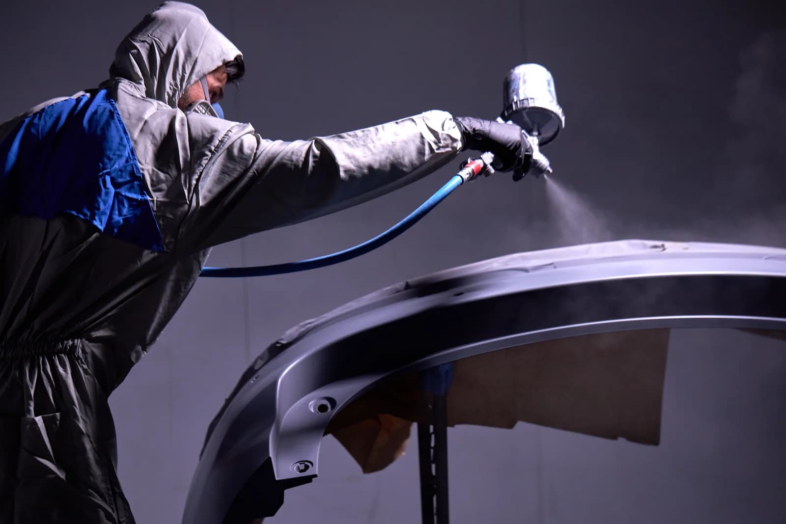 Certified painter in protective gear spray-painting a vehicle panel in a professional paint booth