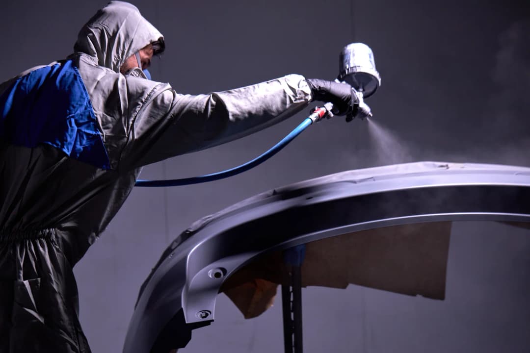 Certified painter in protective gear spray-painting a vehicle panel in a professional paint booth