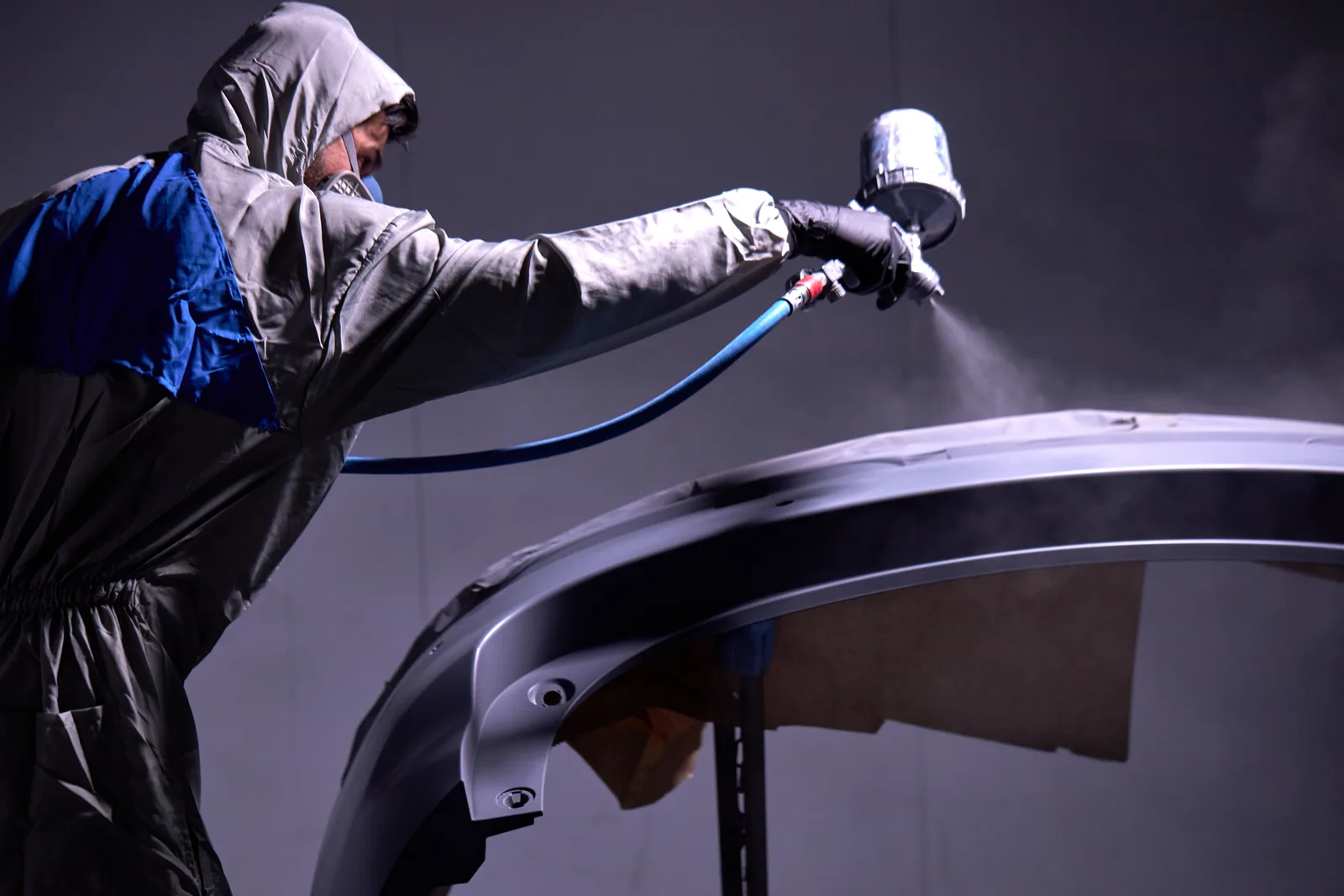 Certified painter in protective gear spray-painting a vehicle panel in a professional paint booth